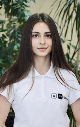Жінка з темним волоссям у футболці, світла шкіра, доброзичлива. У приміщенні, позаду видно рослини.